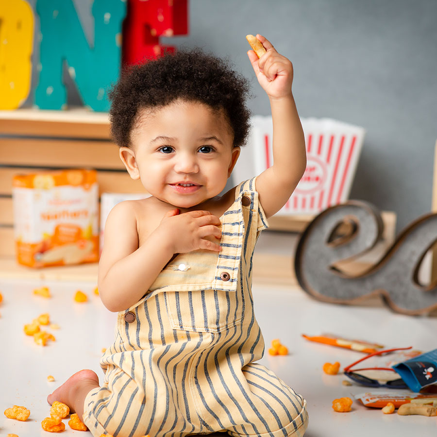 little boy eating cheese puffs with arm in air during first birthday shoot